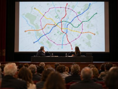 Cittadini a un incontro pubblico in un teatro di quartiere a Milano davanti alla mappa proiettata della nuova metro M6